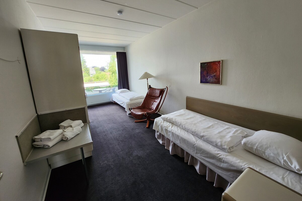 A hotel room with two single beds, a chair, a desk with towels, a lamp, and a painting on the wall.
