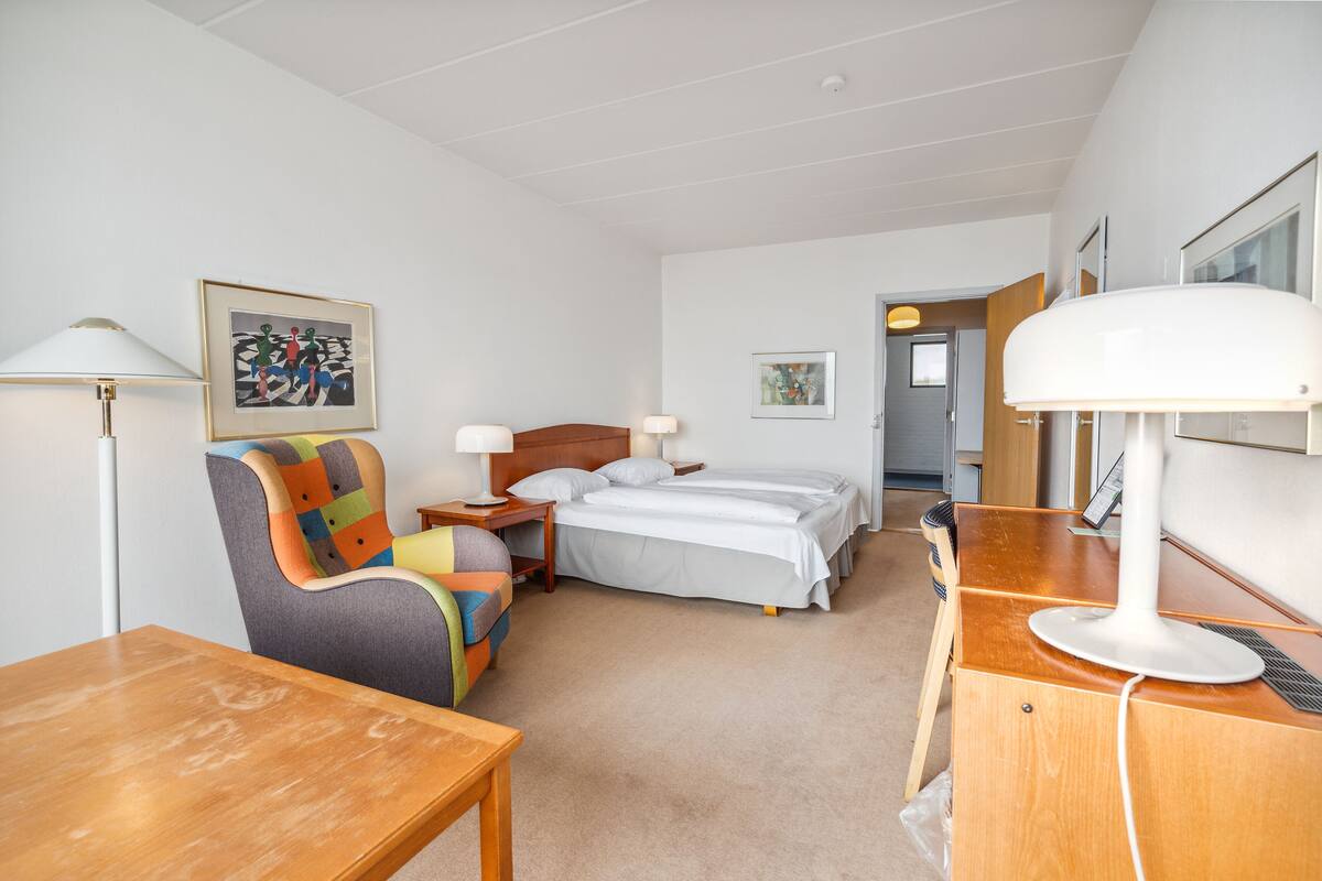 Una habitación de hotel con una cama, un escritorio con computadora, una silla, una lámpara y un cuadro en la pared.