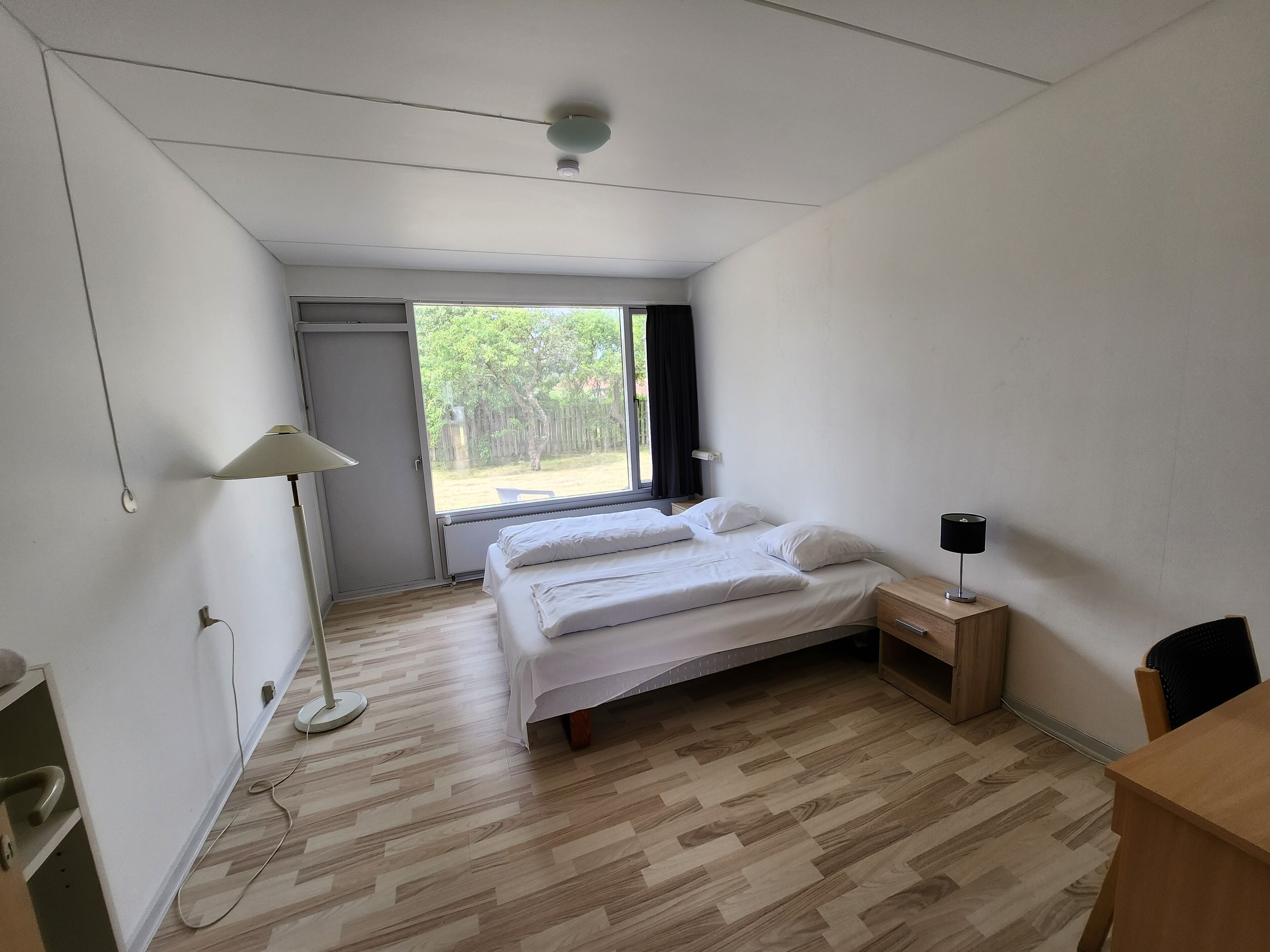 Ein Schlafzimmer mit Bett, Nachttisch, Lampe und Blick ins Freie.