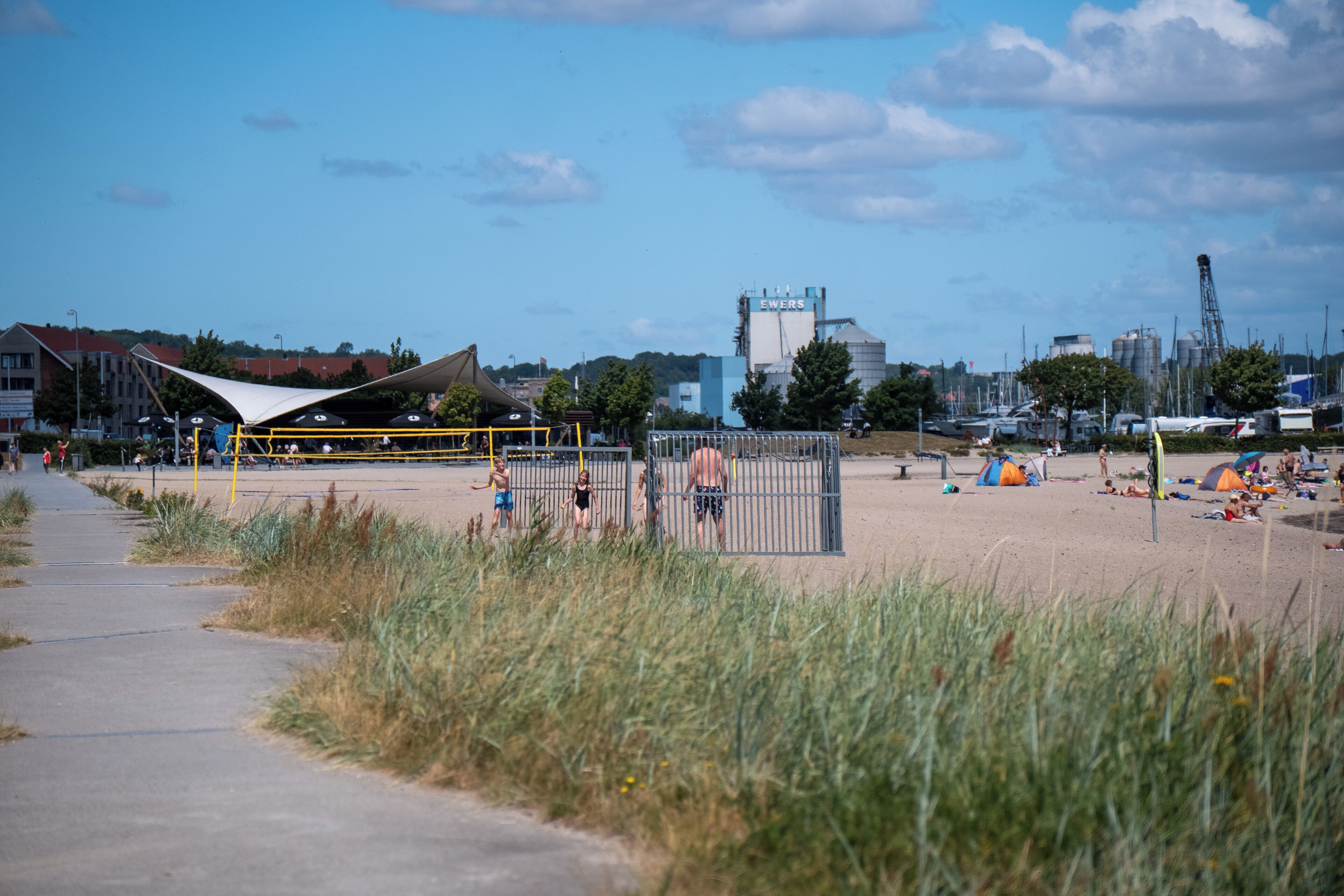 A beach with a fenced area, a playground, and industrial buildings in the background.