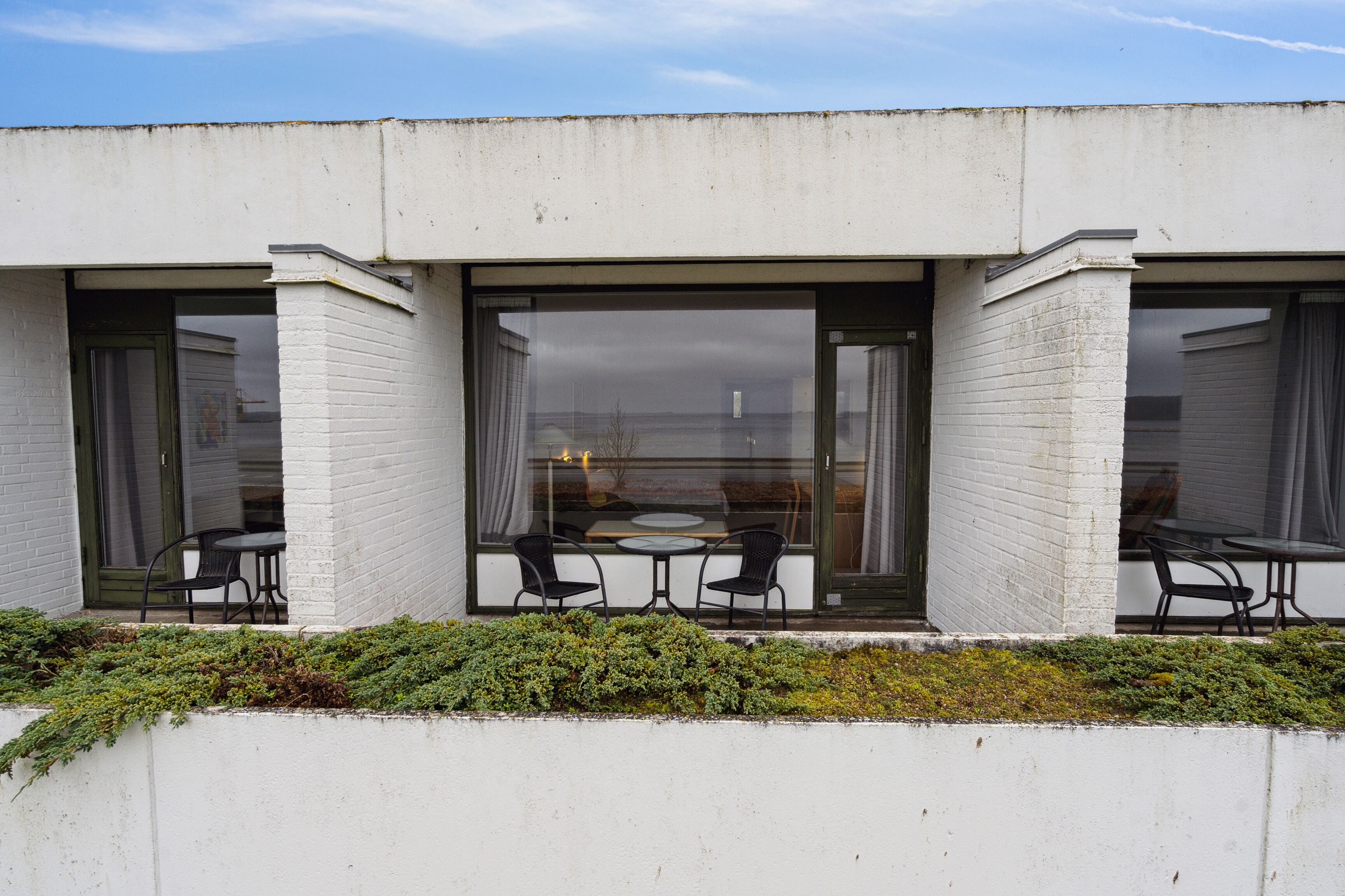 A modern building with large glass doors and windows, a white exterior, and a small outdoor seating area with two tables and four chairs.