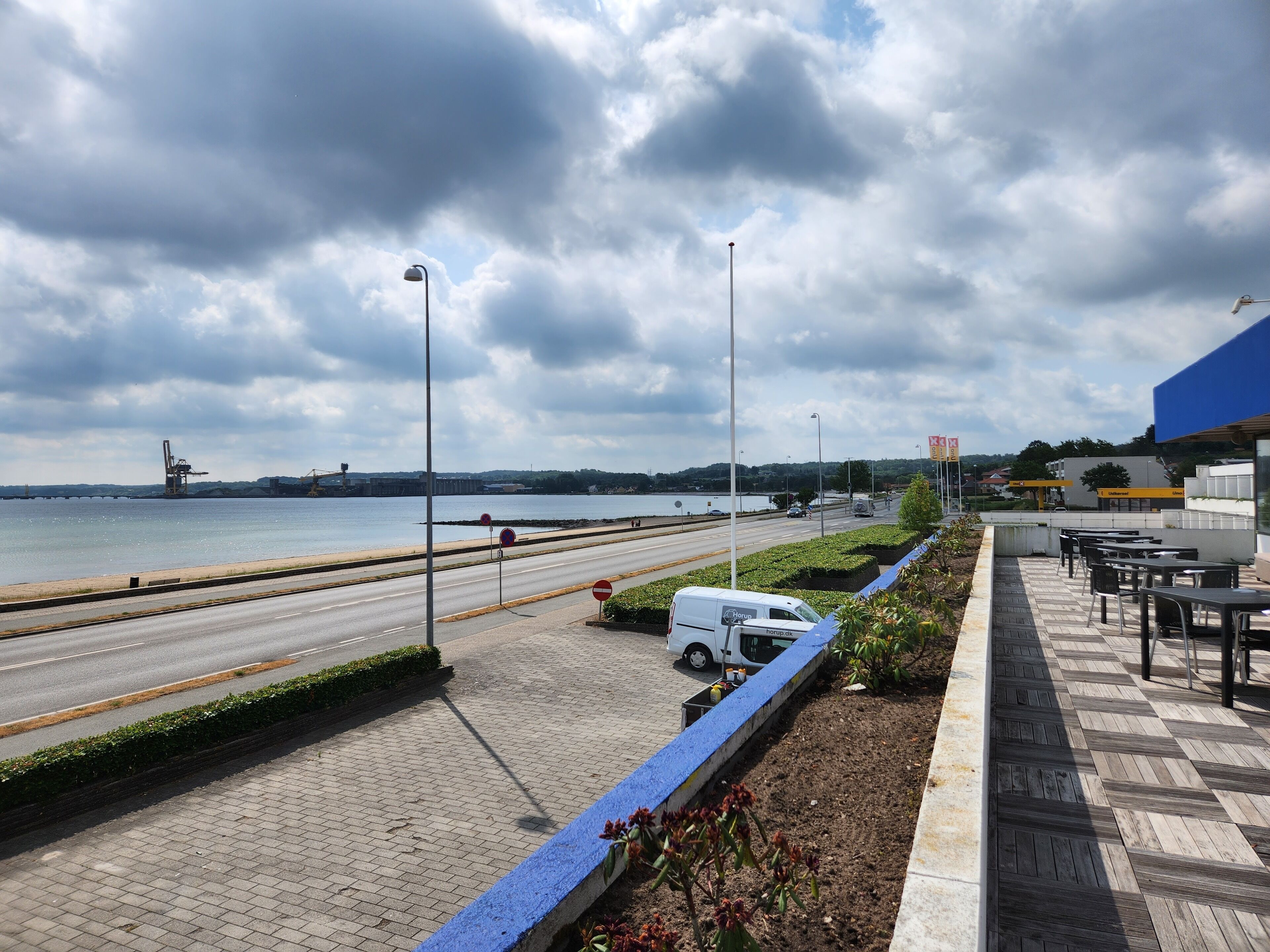 Une promenade en bord de mer avec un chemin piétonnier pavé, un fourgon blanc garé au bord de la route et, au loin, un bâtiment surmonté d’un toit bleu.