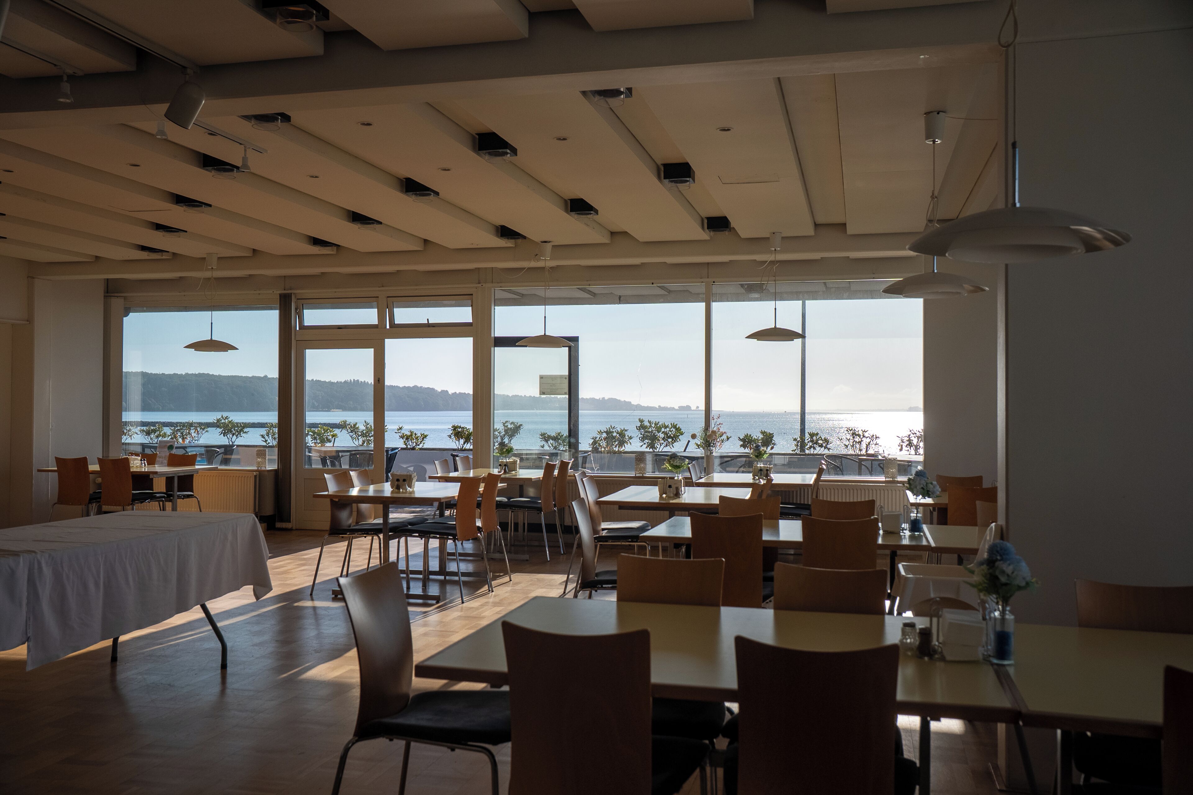 Un restaurante con amplios ventanales que ofrecen vista al mar. Mesas y sillas dispuestas para comer.