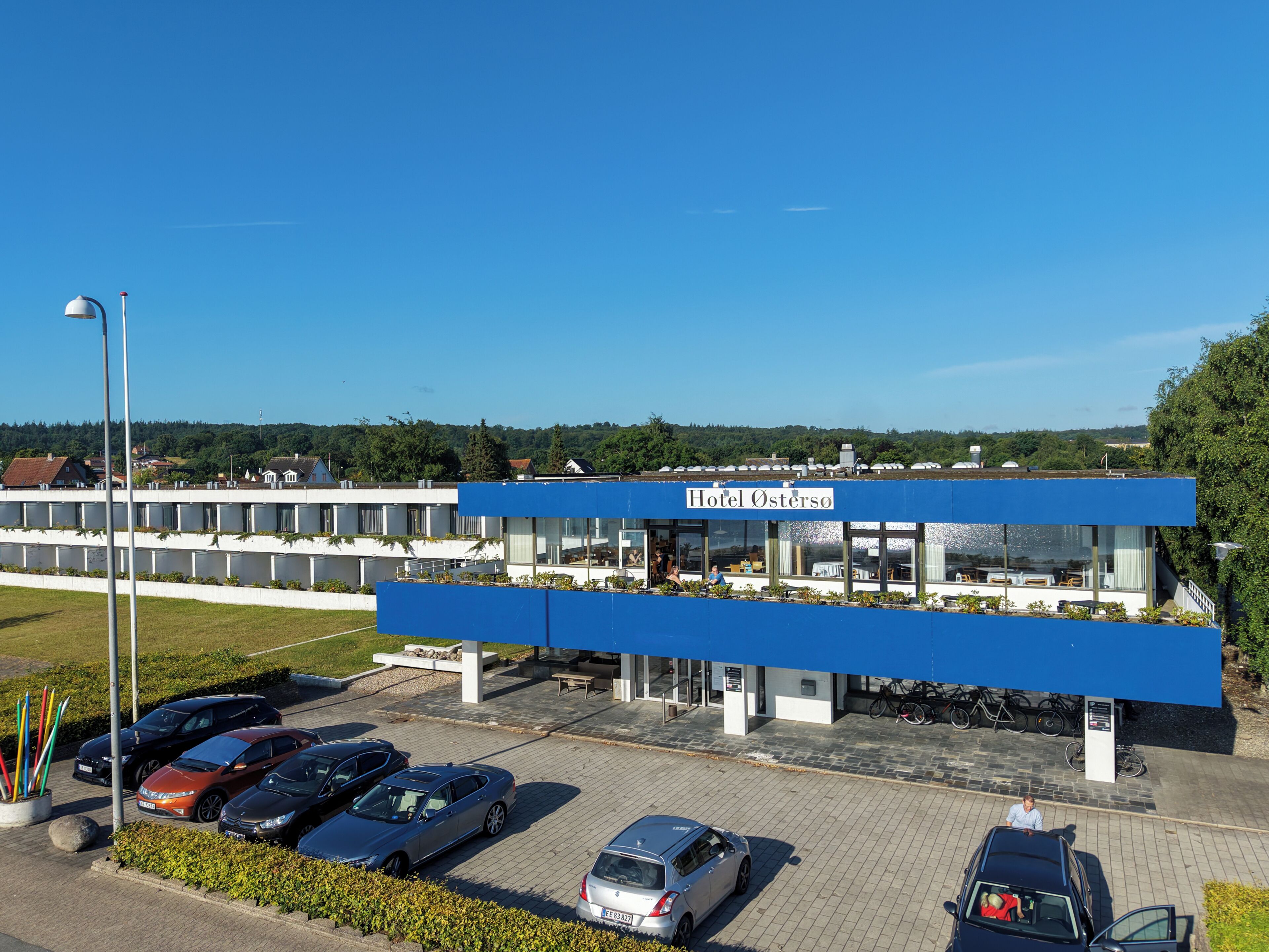 A hotel with a blue facade, a parking lot with several cars, and a clear sky.