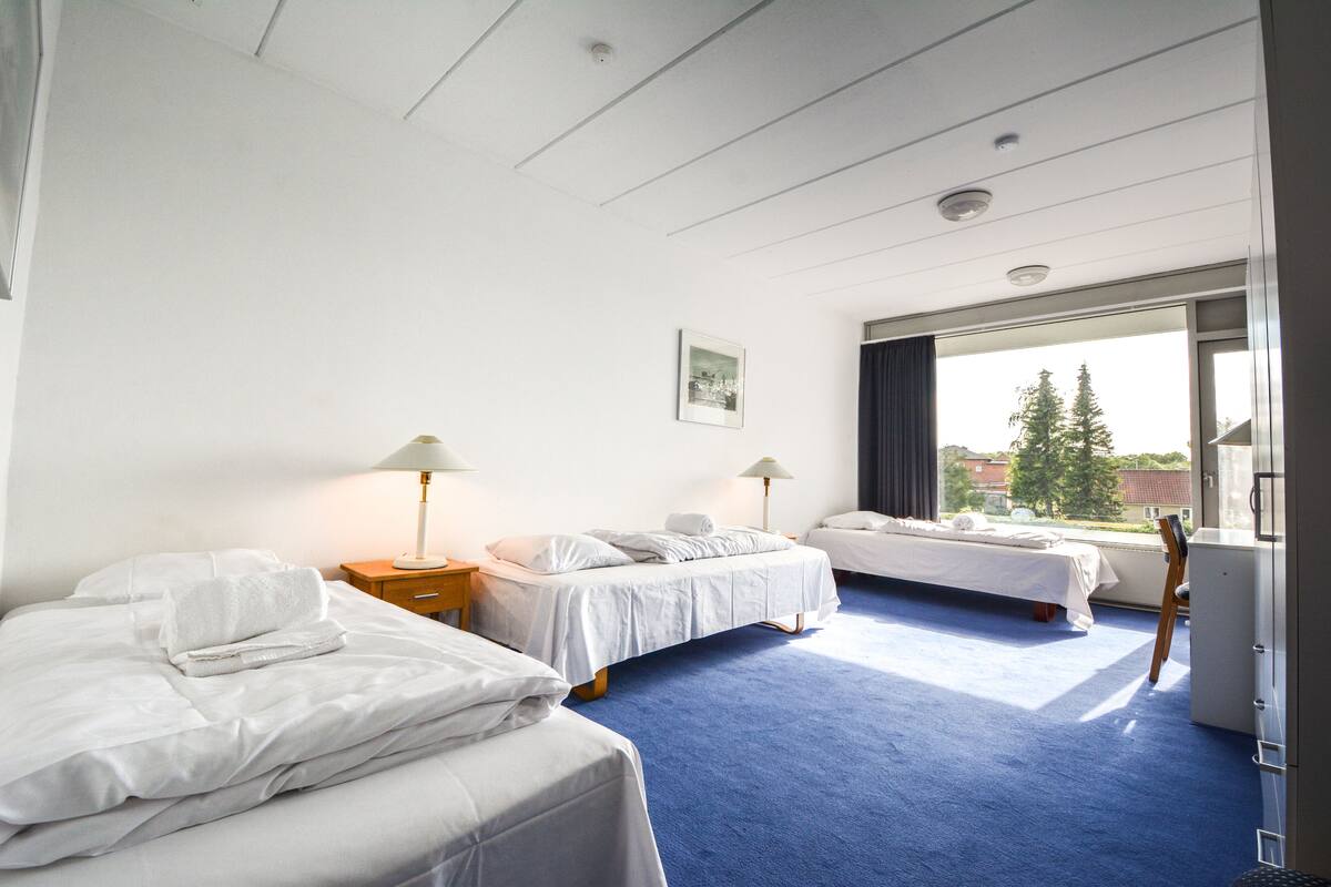 Habitación de hotel con dos camas, un escritorio, una silla y un amplio ventanal con vistas a árboles y edificios.