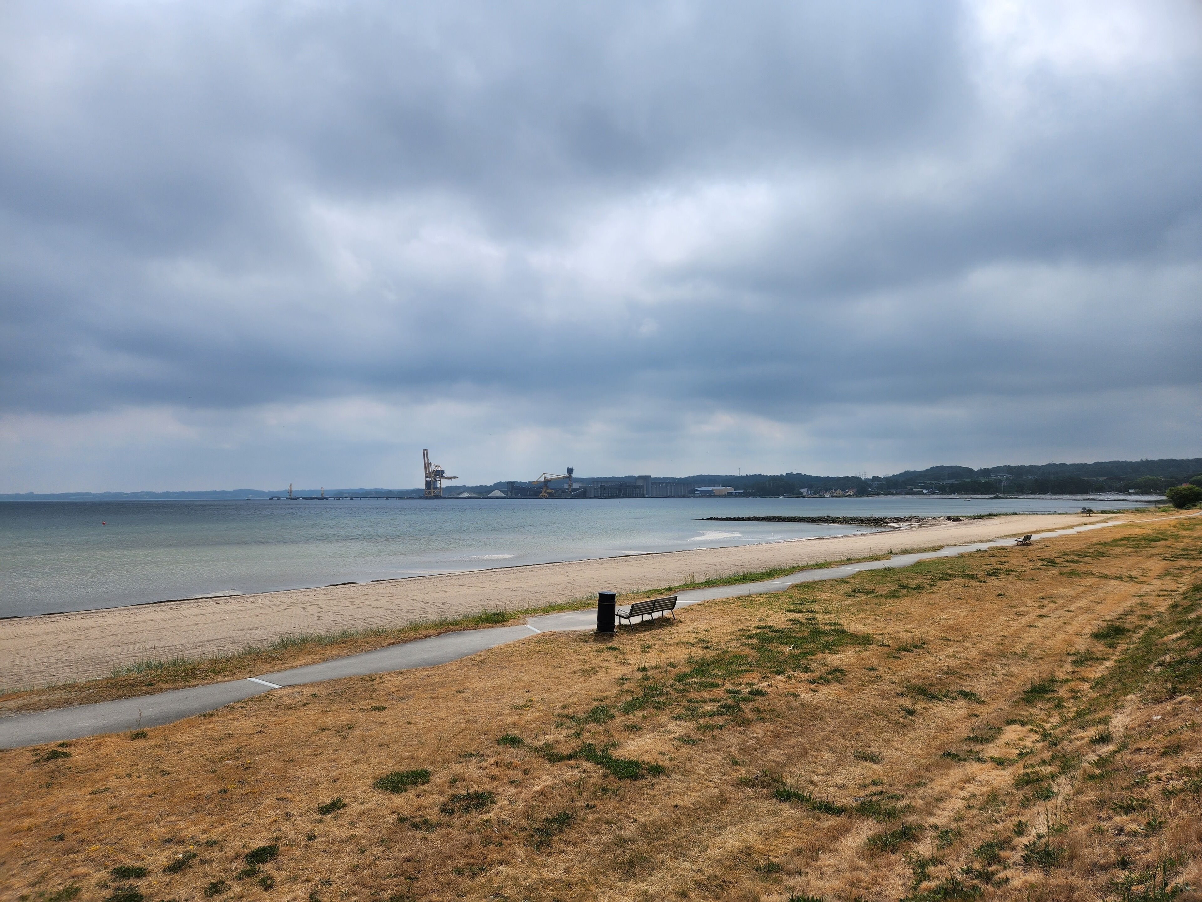 Ein Strand mit einer Bank, einem Pfad und Industriegebäuden im Hintergrund.