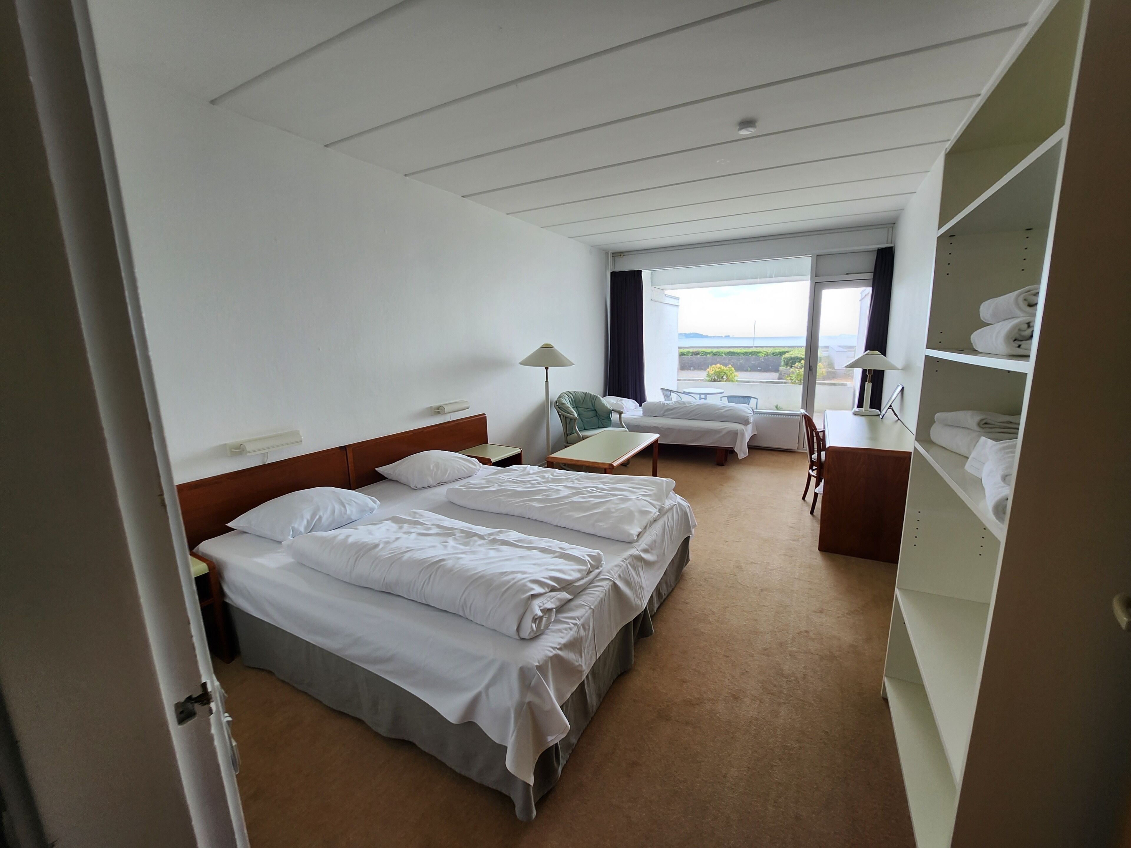 A hotel room with two beds, a desk, a chair, a lamp, and a view of the outdoors.