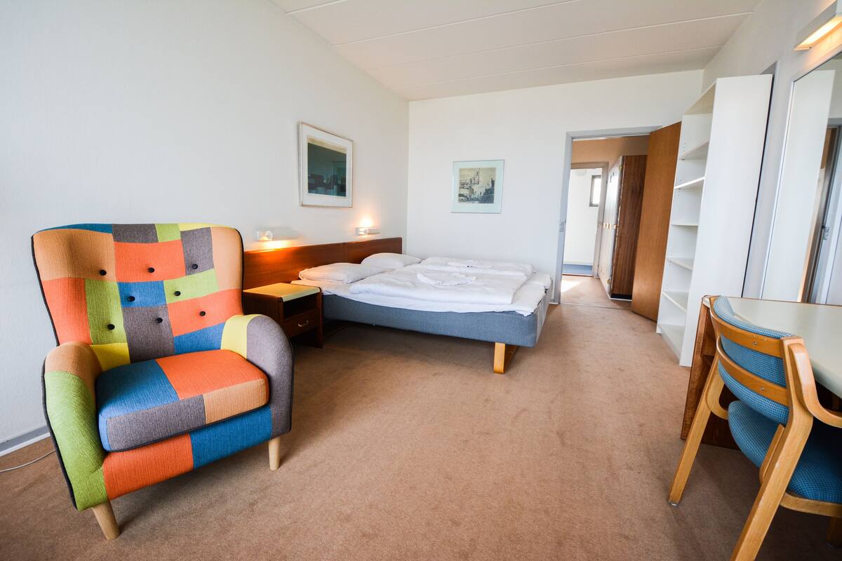 Habitación de hotel con un sillón colorido, una cama con sábanas blancas, una mesita de noche de madera y un escritorio con silla.