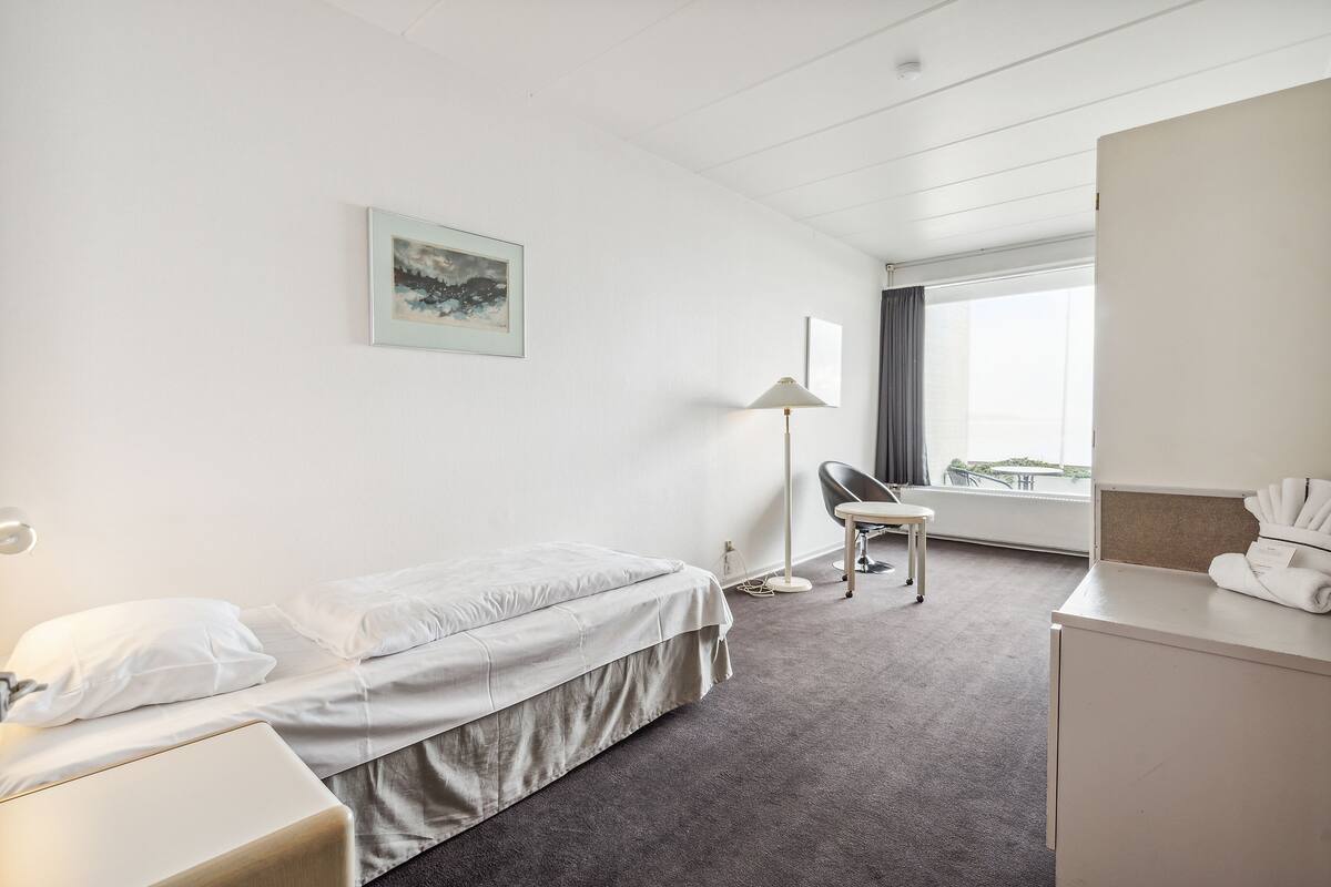 Habitación de hotel con cama, escritorio, silla, lámpara y ventana con cortinas.