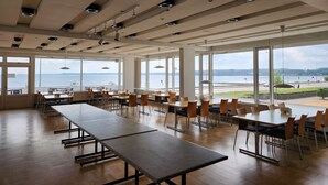 Un amplio comedor con mesas y sillas largas, ventanas grandes que ofrecen vistas a un cuerpo de agua y un paisaje urbano distante.