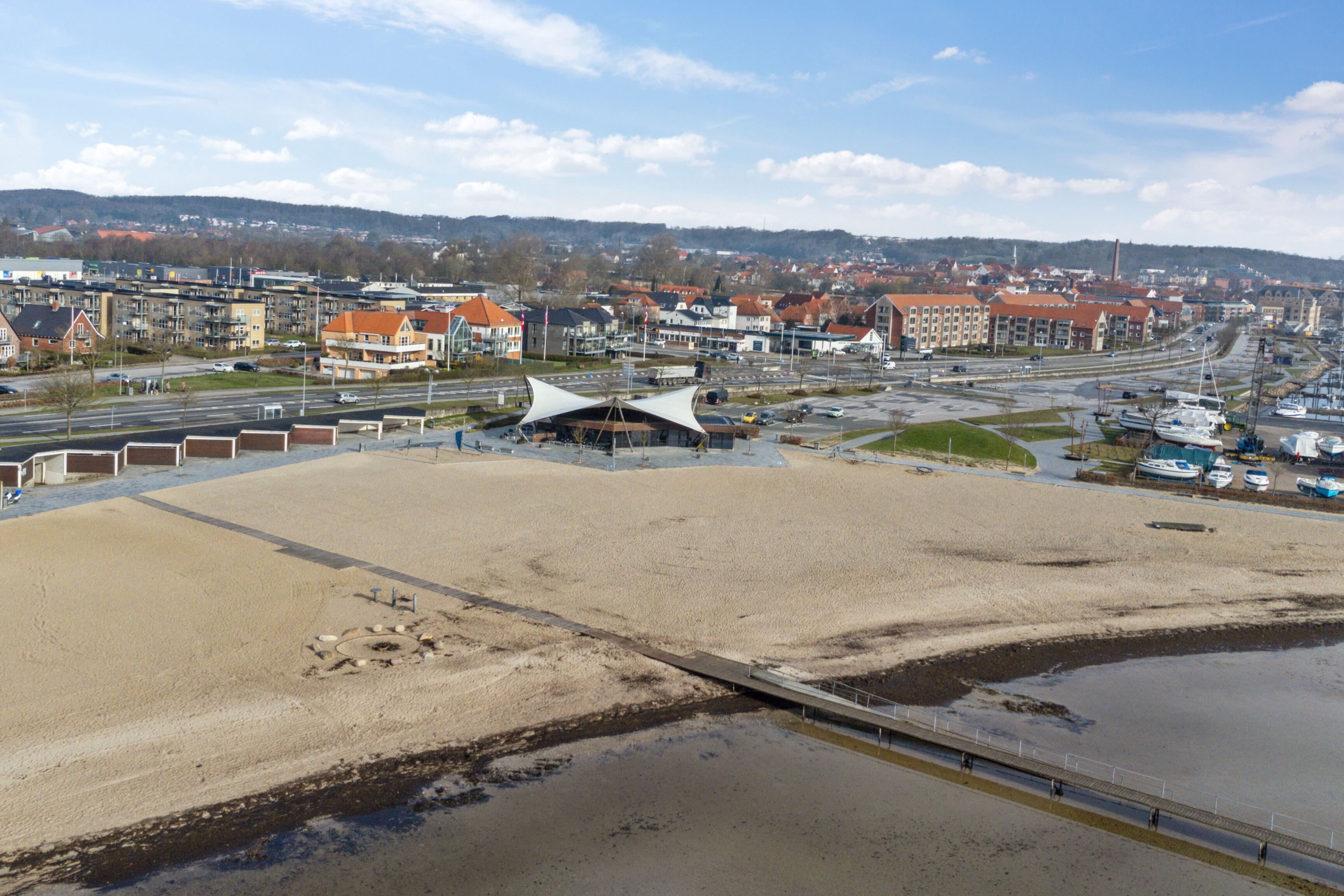 Et kystområde med en sandstrand, en moderne bygning med et unikt tag og en lystbådehavn med både.