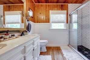 Een badkamer met een houten plafond, witte wanden, een wit toilet, een douchecabine van glas en een witte vloerkleed.