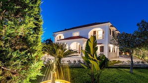 A well-maintained garden with a fountain, lush greenery, and a large white building with multiple arches and balconies. - Aigli Villa (Zakynthos)
