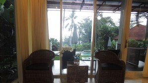 A room with two wicker chairs, a small table, and a view of a pool and palm trees through large glass doors. - Oase Van Java (Gedong)