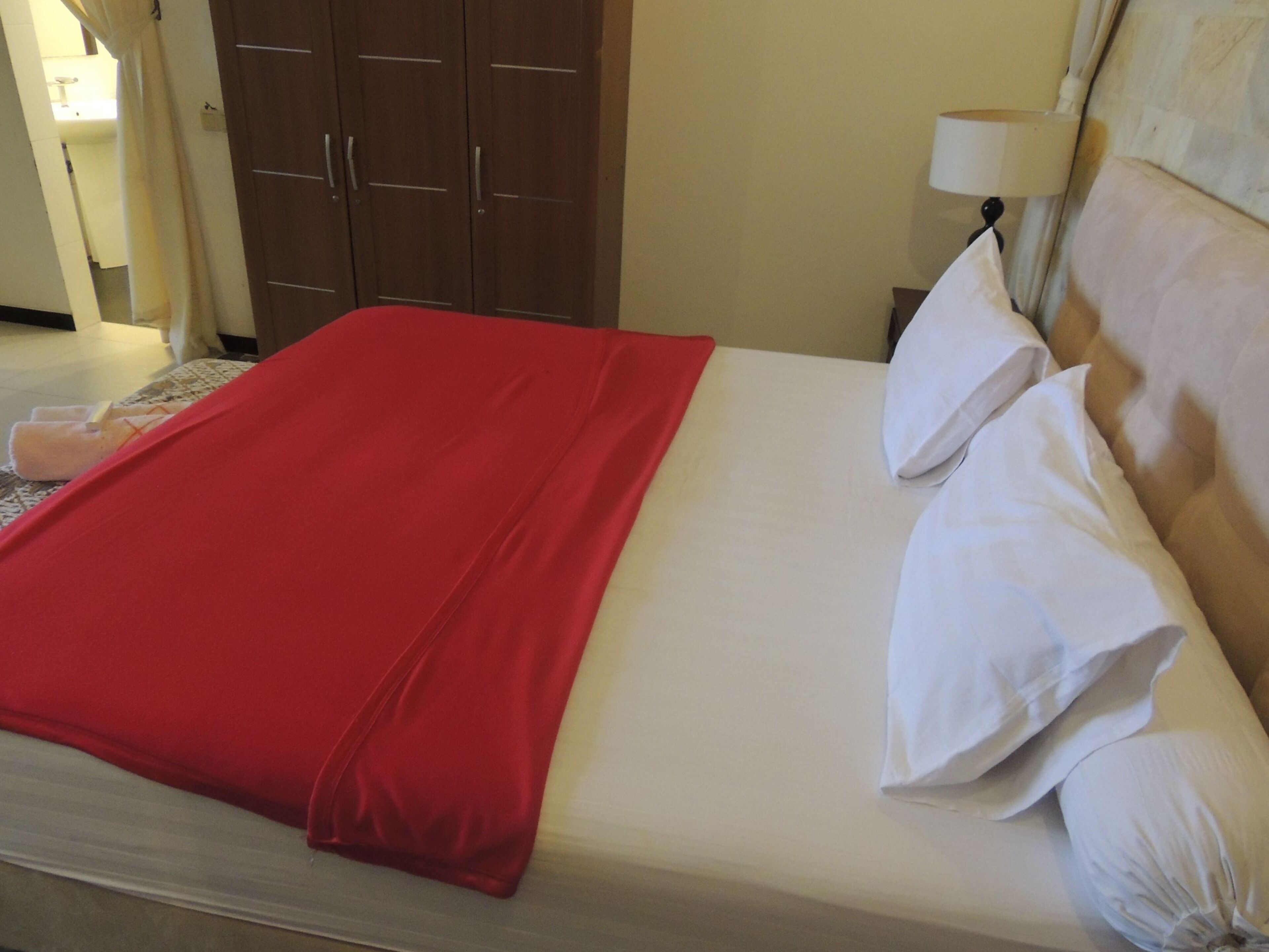 A neatly made bed with a red blanket and white pillows, a wooden wardrobe, and a lamp on the nightstand.