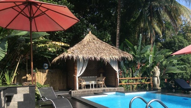 A swimming pool with a thatched-roof hut, lounge chairs, and a red umbrella. - Oase Van Java (Gedong)