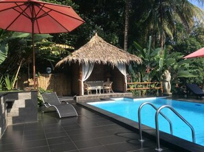 Une piscine avec un petit abri à toit de chaume, des chaises longues et un parasol rouge.