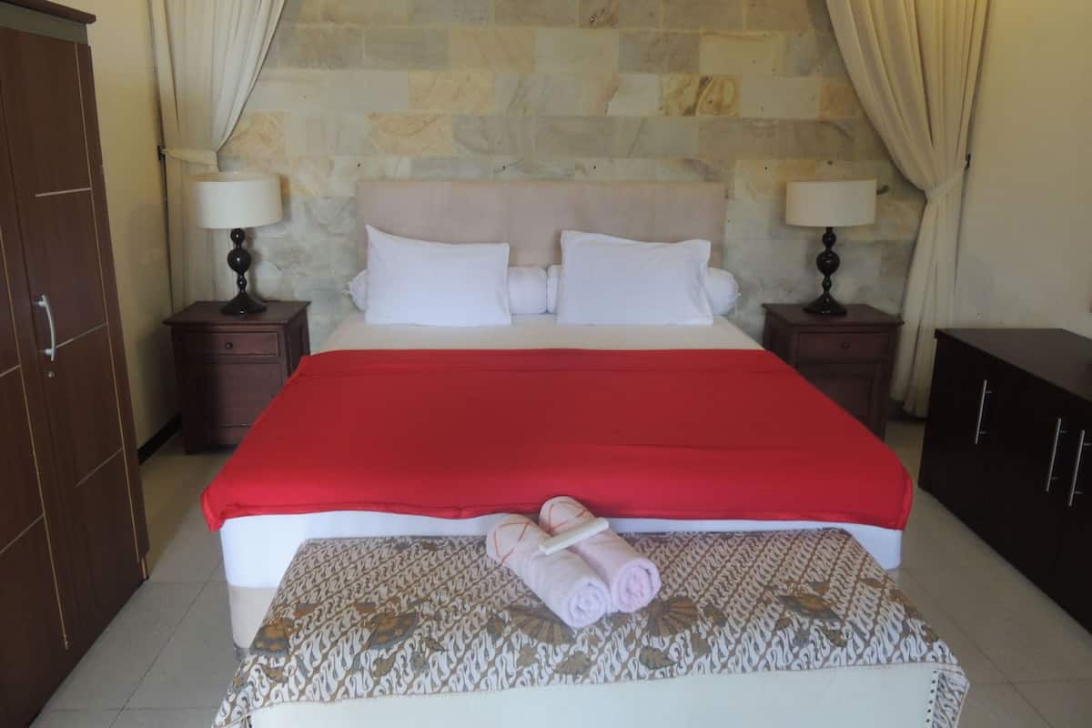 Uma cama bem arrumada com um cobertor vermelho, duas luminárias de cabeceira, um tapete estampado e uma toalha enrolada em um banco.