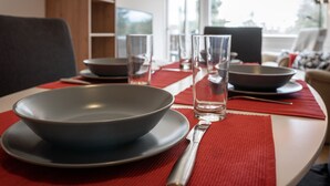 Una mesa de comedor dispuesta con un tazón, platos, vasos y cubiertos, con un mantel individual rojo y vistas al exterior a través de una ventana.