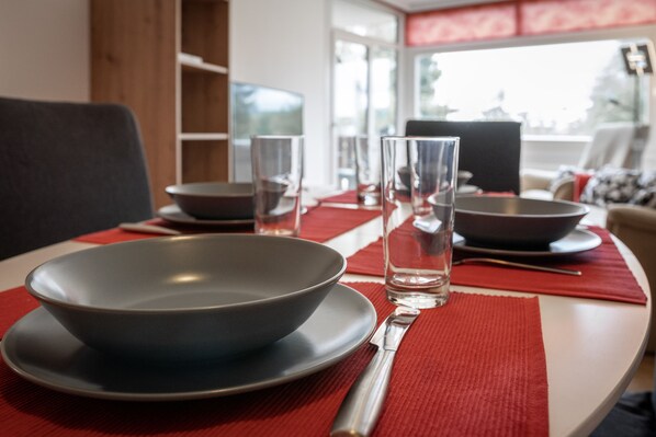 Une table à manger dressée avec un saladier, des assiettes, des verres et des couverts, agrémentée d’un set de table rouge et offrant une vue sur l’extérieur par une fenêtre.