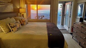 A bedroom with a large bed, a nightstand, a lamp, and a view of the beach at sunset. - Totally Remodeled Beachfront Studio-Step out your door onto the sand. Really! D (Bradenton Beach)