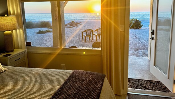 A bedroom with a view of the beach at sunset, a bed, a nightstand, and a lamp. - Totally Remodeled Beachfront Studio-Step out your door onto the sand. Really! D (Bradenton Beach)