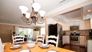 A dining room with a wooden table set for four, white chairs, and a chandelier. Adjacent is a kitchen with stainless steel appliances and white cabinetry.