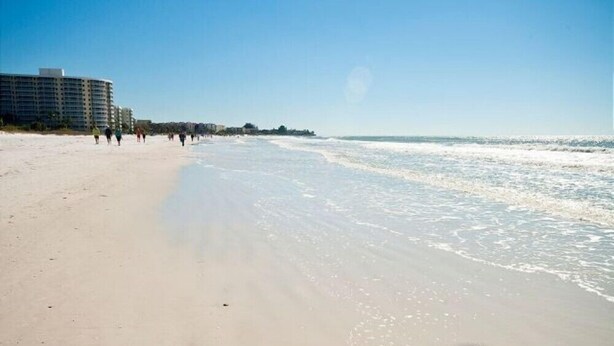 A beach with a long stretch of sand and a few people walking. There are buildings in the background.