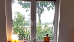 Ein gemütliches Wohnzimmer mit einem Ecksofa, einem kleinen Holztisch mit Dominosteinen, einem Korbstuhl und einem Fenster mit Blick auf Grünflächen.