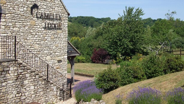 A stone building labeled "CAMILLY LODGE" with a staircase and a view of a lush garden with purple flowers. - In Somerset (Bristol)
