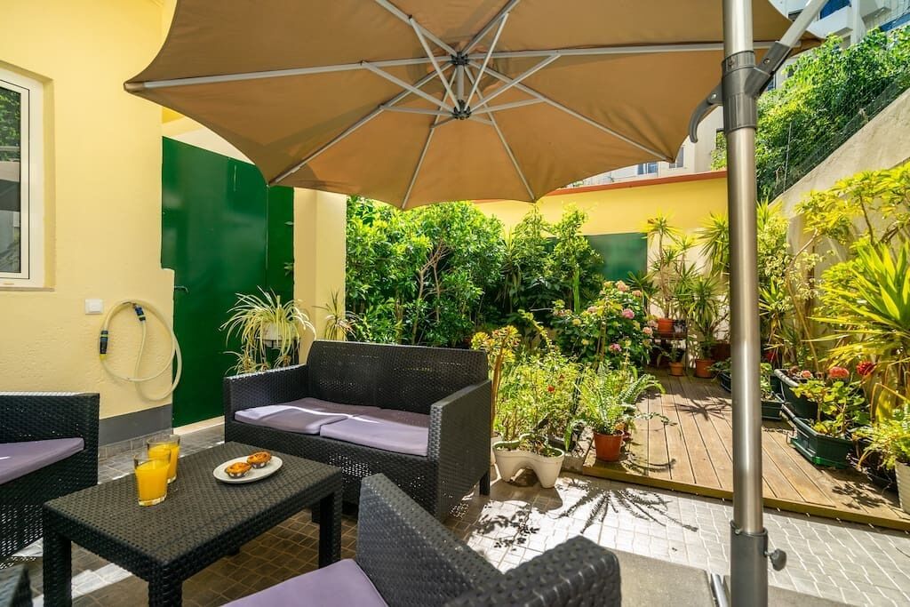 An outdoor patio area with wicker furniture, a table with a plate of food and drinks, and a large umbrella providing shade.