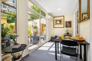 A dining area with a table set for two, a chair, and a small side table with a plant. There are two framed pictures on the wall, and a window with a view of greenery outside.