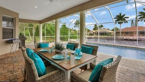 A covered outdoor dining area with wicker furniture and teal cushions, overlooking a swimming pool and palm trees.