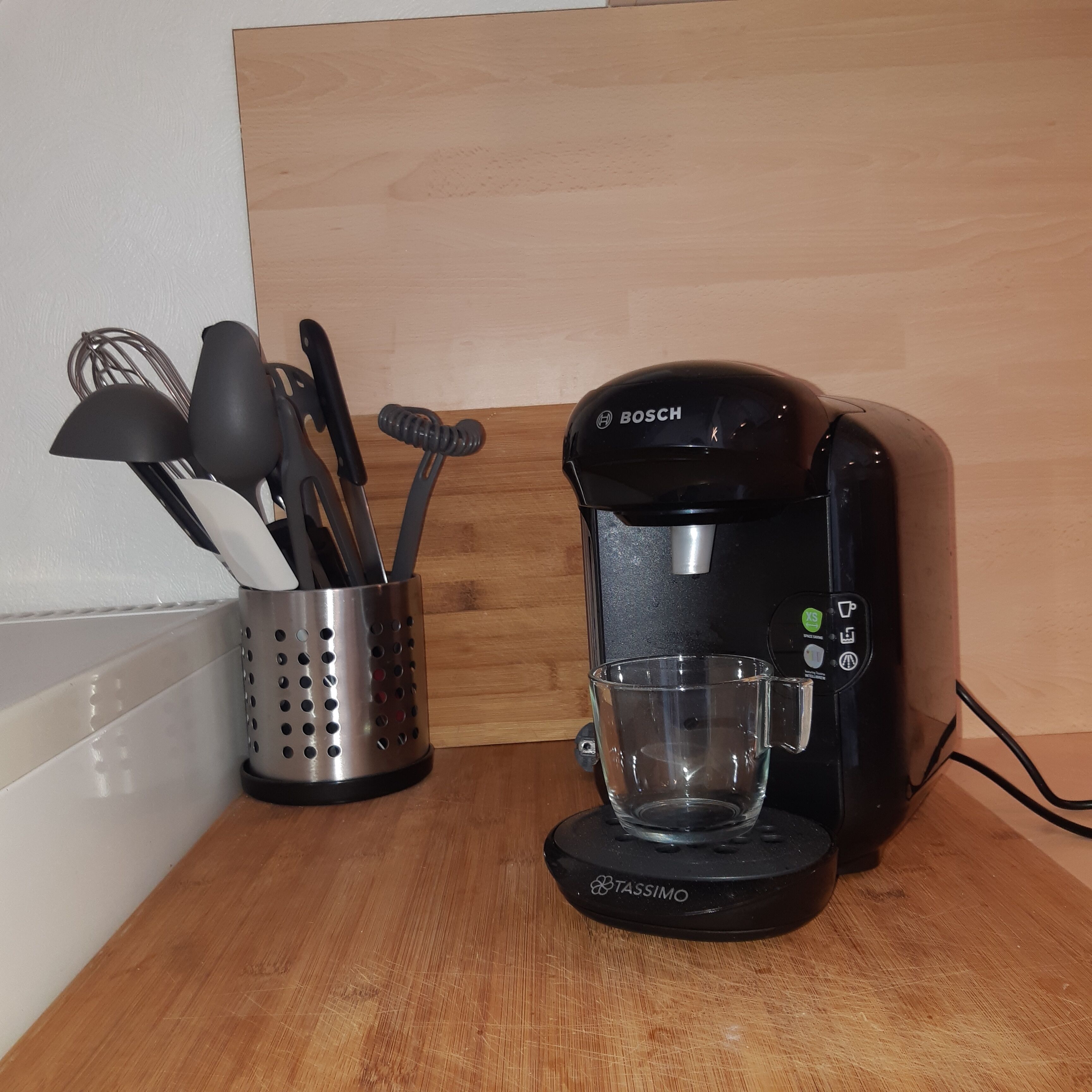 Eine Kaffeemaschine mit einer transparenten Glaskaraffe auf einer Holztischplatte, neben einem Besteckhalter.