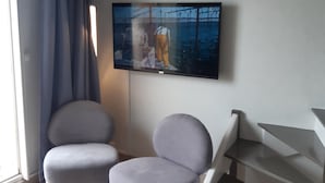 A modern living room with a flat-screen TV mounted on the wall, two grey armchairs, a staircase, and a large window with curtains. - Beachfront apartment on Juno Beach (Bernières-sur-Mer)