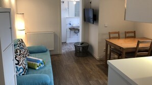 Een gezellig, ruimtelijk ingerichte leefruimte met een bank, een klein rond tafeltje, een houten eettafel met stoelen, een aan de muur bevestigde televisie en een zichtbare badkamer op de achtergrond.
