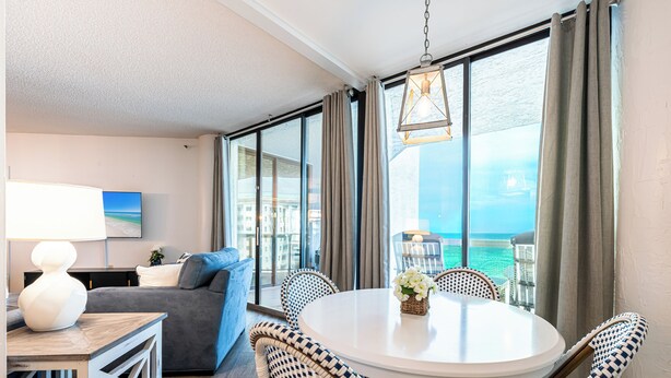 Quarto de hotel com varanda, mesa de jantar redonda, cadeiras de vime, um sofá e vista para o oceano.