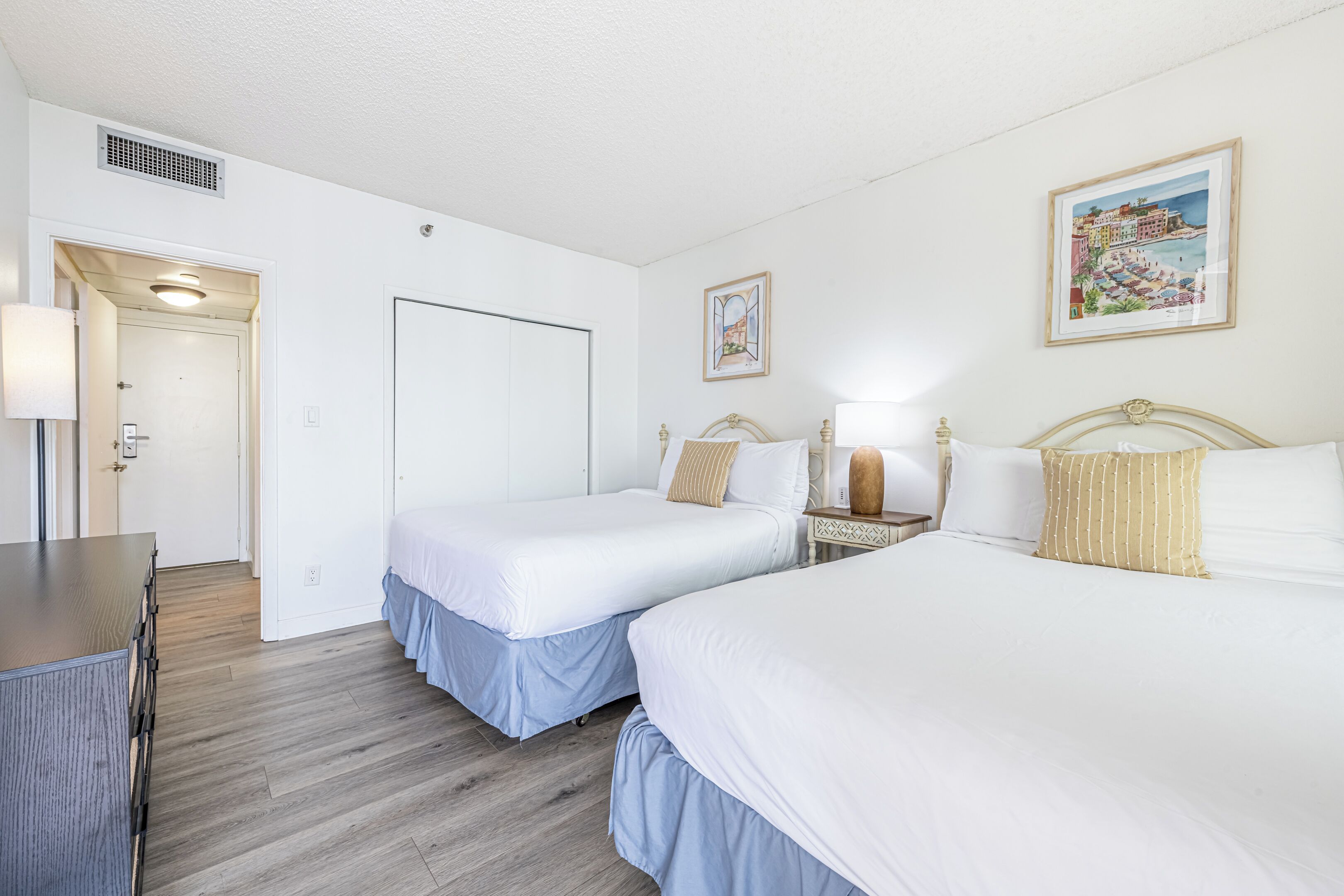 A hotel room with two beds, a desk, a chair, a lamp, and a framed picture on the wall.