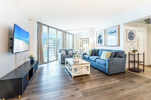 Un soggiorno moderno con divano, tavolino e televisione. Grandi finestre offrono vista sugli edifici esterni.