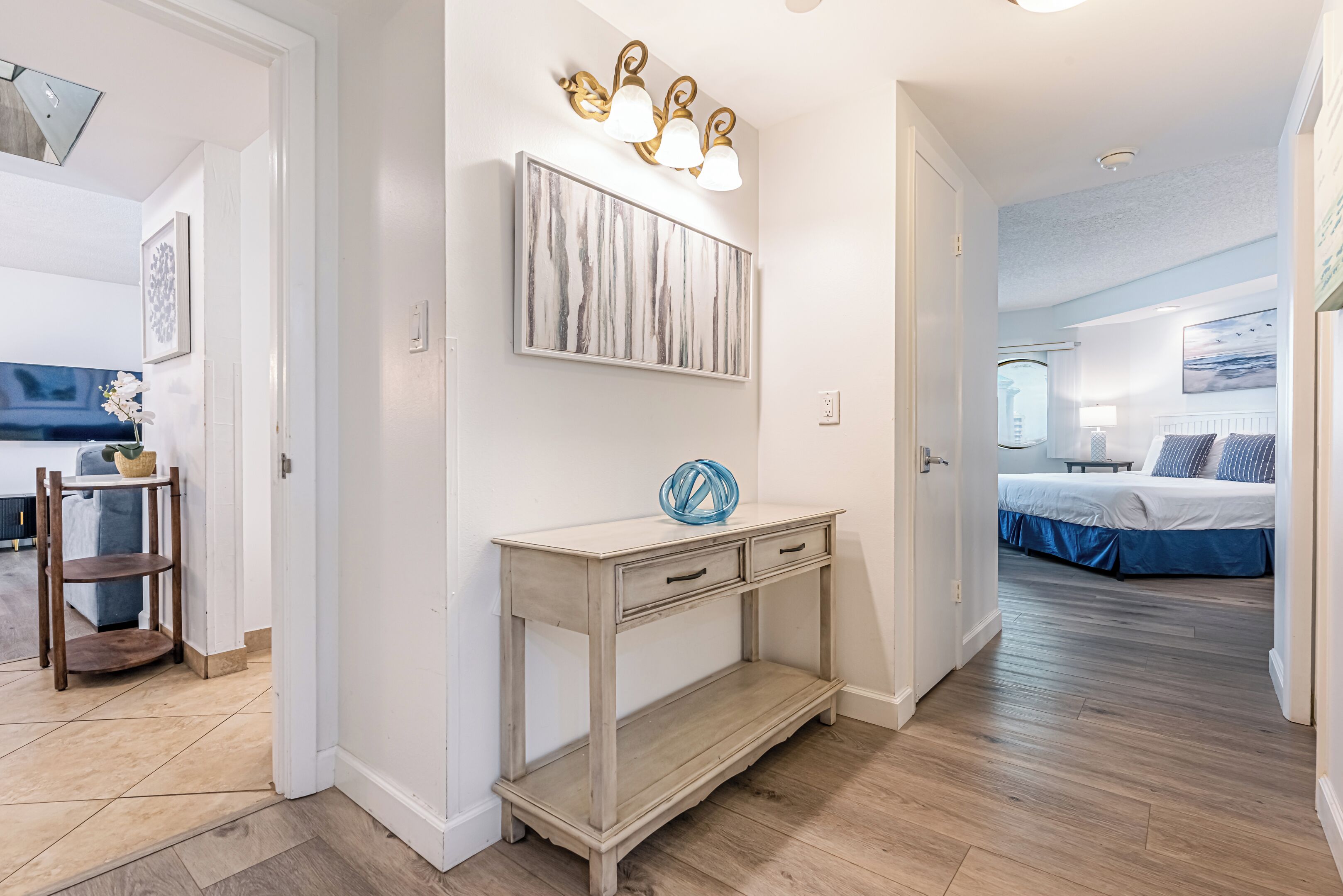 A hallway with a console table, wall art, and a bed in the adjacent room.