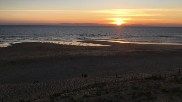Une plage au coucher du soleil, avec une mer calme et quelques personnes qui se promènent.
