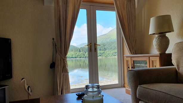 Ein Zimmer mit Blick auf einen See und Berge, ein Holztisch mit einer Glaskaraffe, eine Couch, eine Lampe und ein Fernseher an der Wand.