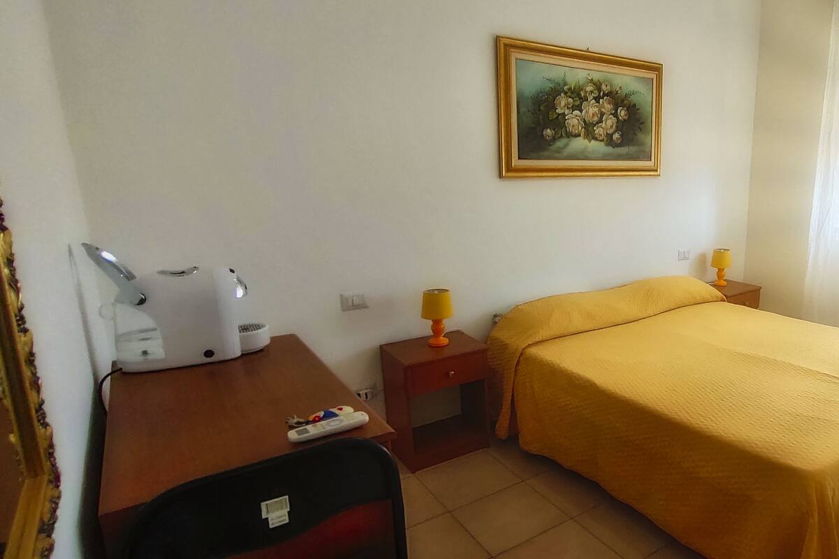A small hotel room with a bed, a desk with a lamp, a chair, and a painting on the wall.