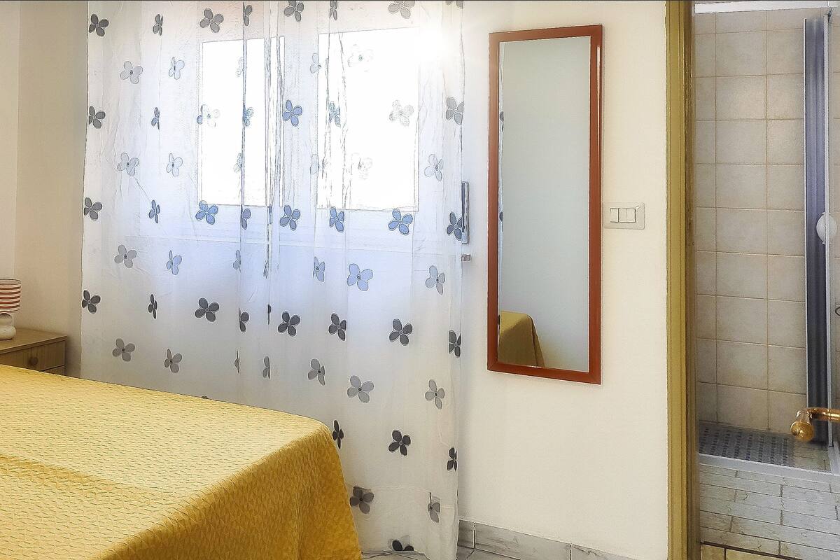 A bedroom with a bed, a window with a floral curtain, a bedside lamp, and a bathroom with a shower.