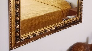 A large ornate mirror with a gold frame reflecting a bed with a yellow bedspread, a framed floral painting, and a bedside lamp. - Hippocampus B&B (Giardini Naxos)