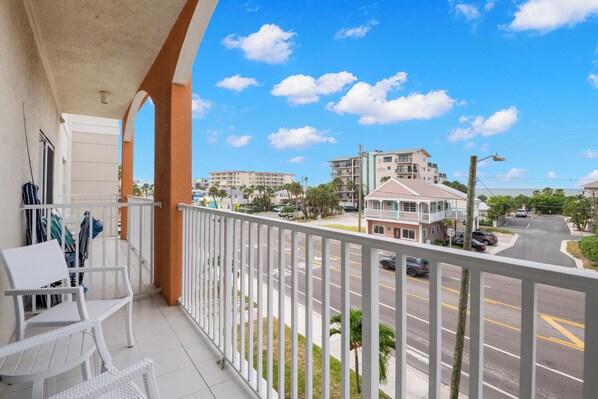 Ein Balkon mit weißem Rattanmöbel, Blick auf eine Straße mit Autos und Gebäuden, und ein klarer, blauer Himmel mit Wolken.