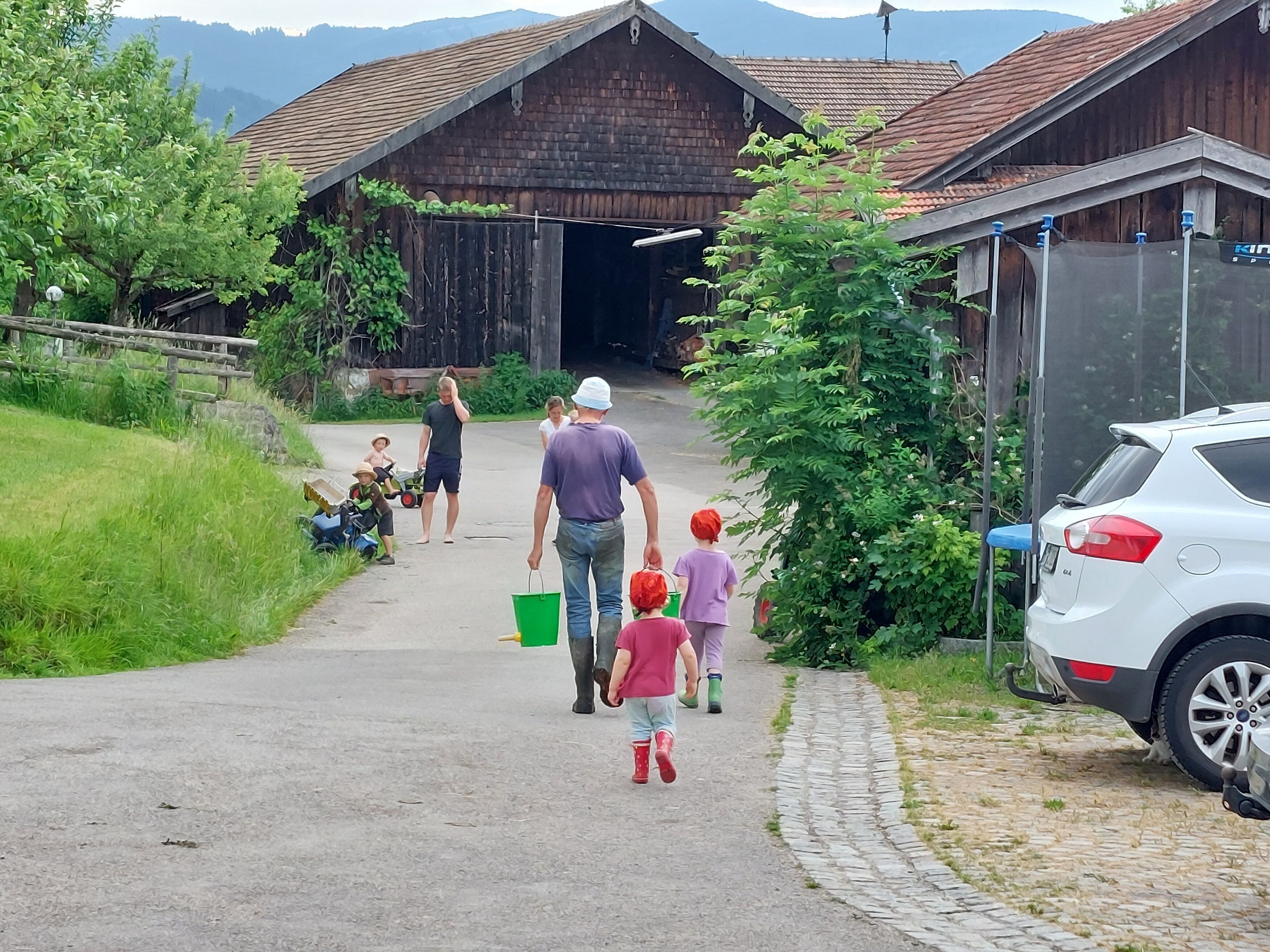 Ein gepflasterter Weg führt zu einem rustikalen Holzhaus mit geneigtem Dach, umgeben von Grünflächen und einem parkenden Auto.