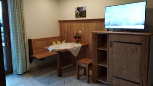 Une pièce chaleureuse avec un banc en bois, une petite table recouverte d’une nappe, un téléviseur posé sur un meuble en bois et un tableau accroché au mur.