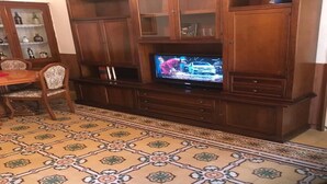 Een houten tv-meubel met een televisie, een kamerbreed tapijt met dessin, en een eethoek met stoelen en een tafel.