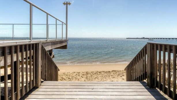 Ein hölzerner Steg führt zu einem Strand mit freiem Blick auf das Meer und einem Pier im Hintergrund.